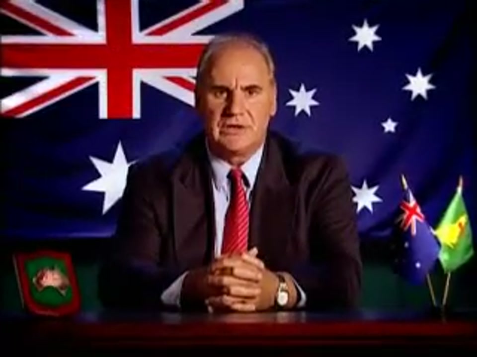 Un-Australianism (2005), scene 1 at 00:20: The scene is a single, continuous shot of Sam Kekovich, a middle-aged white man in a suit and tie, sitting at a large wooden desk. He is positioned as a head of state or news anchor.