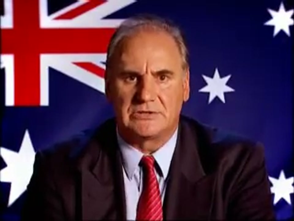 Un-Australianism (2005), scene 4 at 01:21: Kekovich's expression is now one of firm, resolute instruction. He is giving a direct command to the nation. The shot remains fixed. At the very end, white text appears over the lower third of the screen.