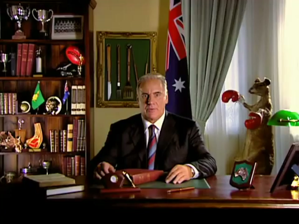 Address to the Nation (2006), scene 1 at 00:05: The scene opens on Sam Kekovich, a middle-aged man in a dark suit, seated at a large, polished wooden desk.