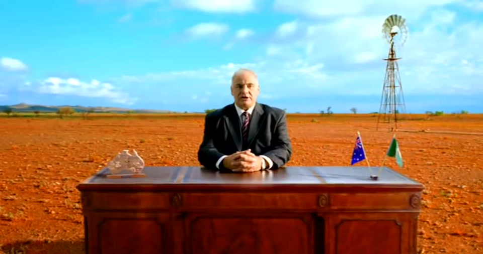 Australia Day Address (2008), scene 1 at 00:06: Sam Kekovich, a middle-aged man in a dark suit and tie, sits at a large, formal wooden desk. He looks directly at the camera with a serious, resolute expression.