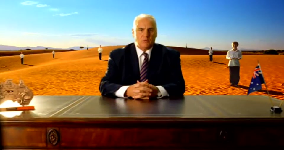 Australia Day Address (2008), scene 4 at 00:53: The scene shifts to a vast, undulating desert of golden sand dunes. The "waiters" are positioned on the dunes in the distance. The shot is wider, emphasizing the isolation and grandeur of the setting.