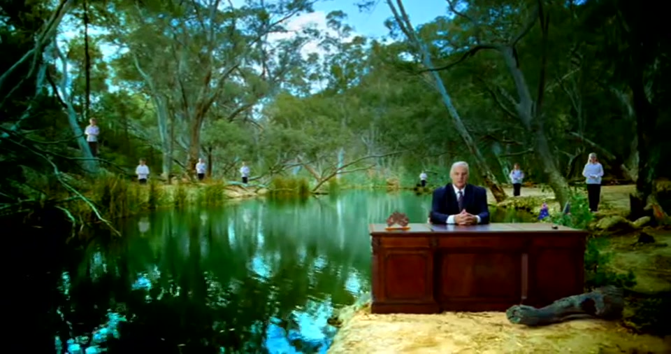 Australia Day Address (2008), scene 5 at 01:00: The desk is now beside a calm river or billabong, surrounded by eucalyptus trees. This is a classic "bush" setting. A kookaburra figurine is now visible on the desk.