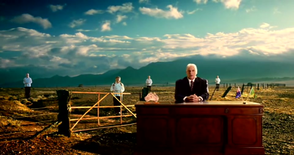 Australia Day Address (2008), scene 6 at 01:19: A montage of increasingly grand and absurd locations: Kekovich's desk is on a dramatic cliff edge overlooking a valley; then behind a farm gate in a rural landscape; finally, the camera pulls back to reveal him and his…