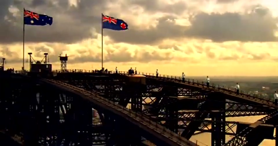 Australia Day Address (2008), scene 6 at 01:25: A montage of increasingly grand and absurd locations: Kekovich's desk is on a dramatic cliff edge overlooking a valley; then behind a farm gate in a rural landscape; finally, the camera pulls back to reveal him and his…