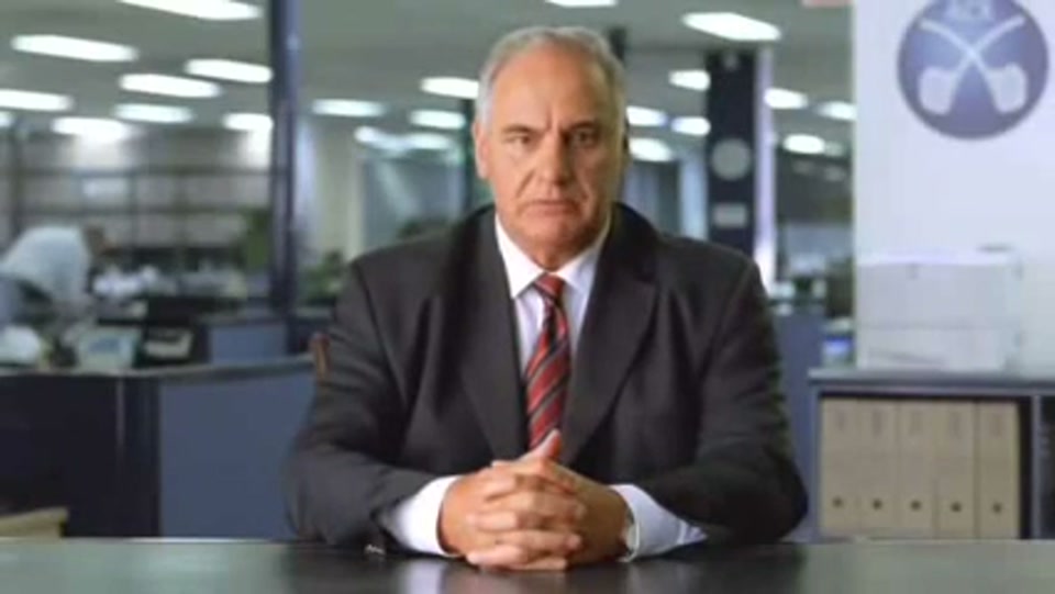 Address to the Nation (GFC Rally) (2009), scene 1 at 00:06: The ad opens with a tight, head-and-shoulders shot of a stern-looking, middle-aged man in a dark suit and red-striped tie, seated at a desk. The setting is a modern, open-plan office.