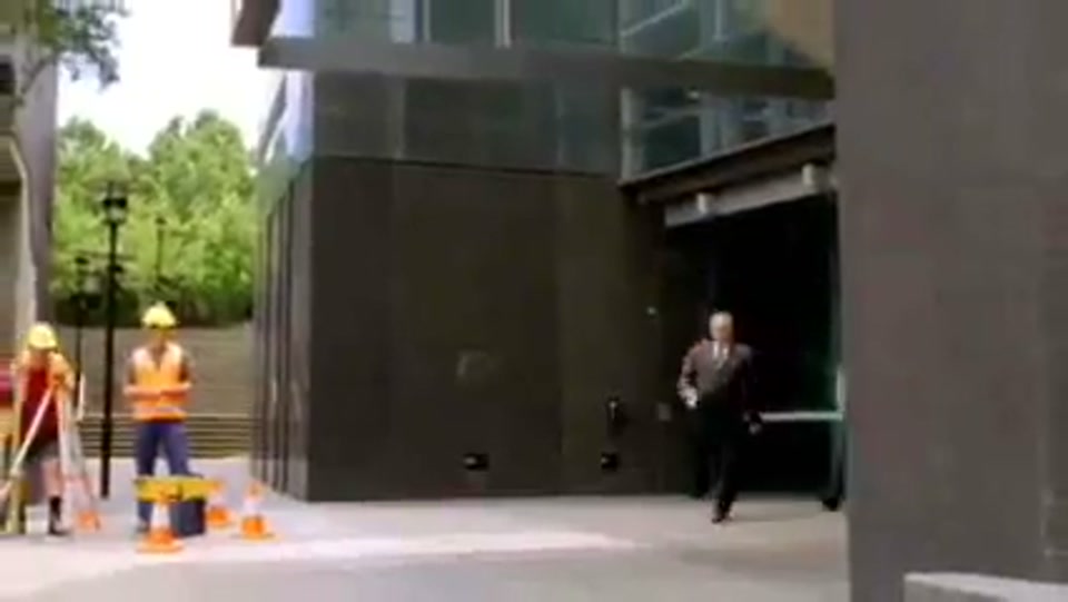 Address to the Nation (GFC Rally) (2009), scene 3 at 00:43: The speaker, now with a small entourage of office workers, walks out of the glass doors of a modern office building.