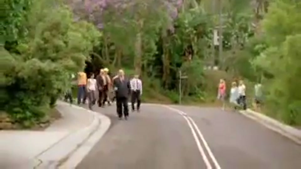 Address to the Nation (GFC Rally) (2009), scene 4 at 00:57: The procession, now a larger crowd, marches away from the city and down a tree-lined suburban road. The followers now include people in casual clothes, jogging outfits, etc.