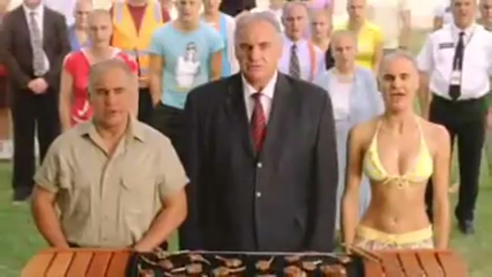 Address to the Nation (GFC Rally) (2009), scene 5 at 01:21: The scene cuts to a vast rally on a grassy area by a beach, with a large bay filled with boats in the background. The speaker stands at a lectern which is actually a barbecue laden with lamb chops.