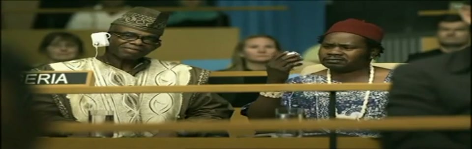 Sam Kekovich UN Speech (2010), scene 3 at 00:30: Kekovich continues his speech, looking directly at the camera. The camera cuts to two men in African traditional dress and then to a man and woman under a "TURKEY" placard, who look on with stern, unamused expressions.