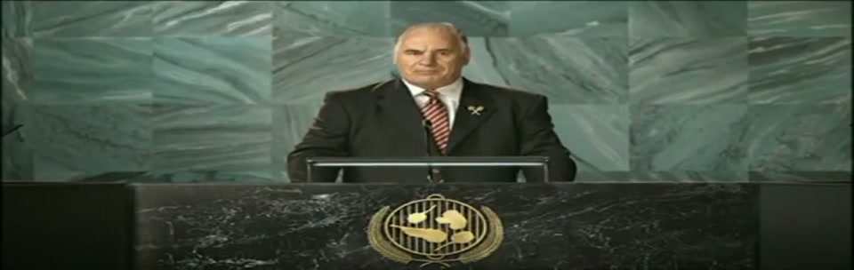 Sam Kekovich UN Speech (2010), scene 3 at 00:33: Kekovich continues his speech, looking directly at the camera. The camera cuts to two men in African traditional dress and then to a man and woman under a "TURKEY" placard, who look on with stern, unamused expressions.