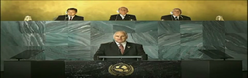 Sam Kekovich UN Speech (2010), scene 3 at 00:26: Kekovich continues his speech, looking directly at the camera. The camera cuts to two men in African traditional dress and then to a man and woman under a "TURKEY" placard, who look on with stern, unamused expressions.