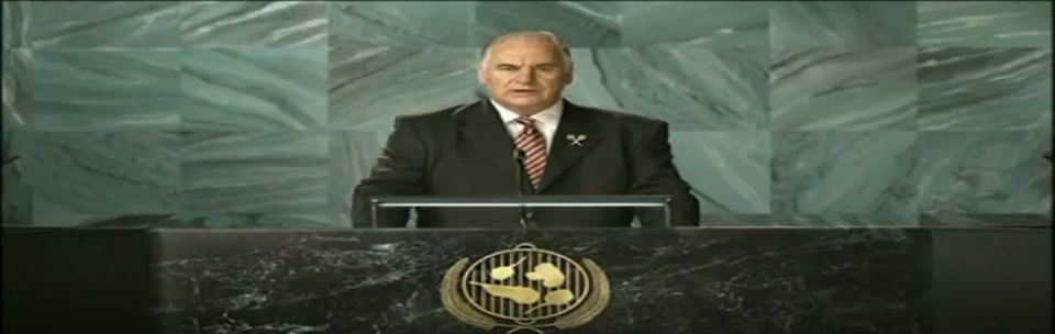 Sam Kekovich UN Speech (2010), scene 4 at 00:55: Kekovich becomes more animated, gesturing as he presents his solution. The camera remains focused on him at the podium, with the BBQ-themed seal prominent.