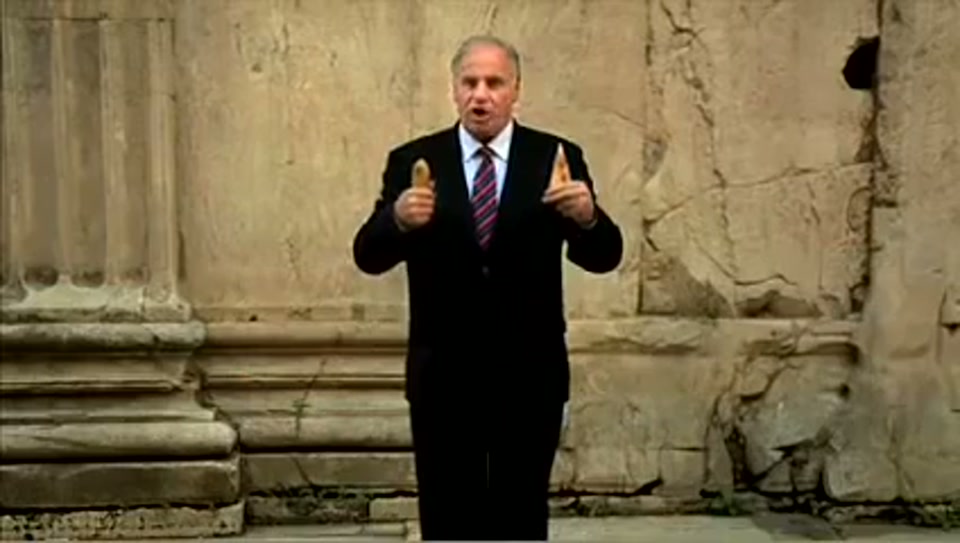 Address to the United Nations of Australia (2011), scene 3 at 00:34: Kekovich stands on a snowy street with a sign pointing towards "Brussels." He then appears in front of the Eiffel Tower, holding up a tiny pair of cooked frog's legs.