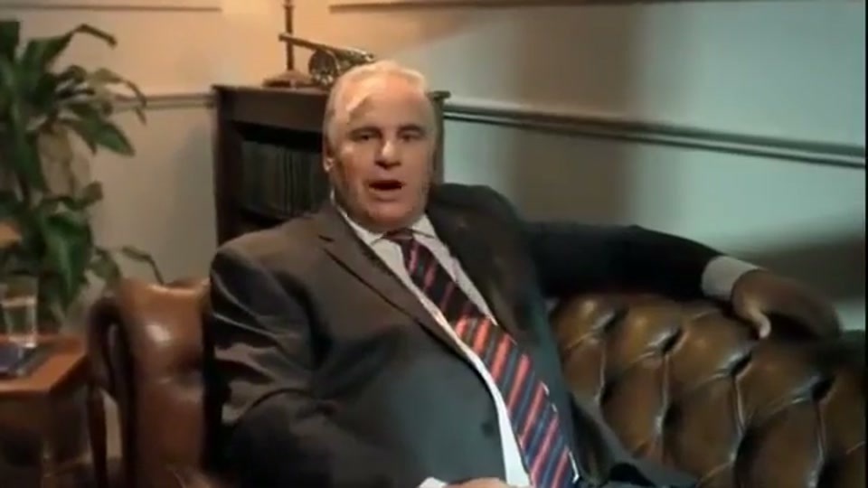 Lambnesia (2013), scene 1 at 00:03: Sam Kekovich, a middle-aged man in a suit, sits slumped on a formal leather chesterfield sofa. The setting is a dimly lit, wood-panelled office, suggesting a seat of power. As he speaks, the camera slowly zooms in.