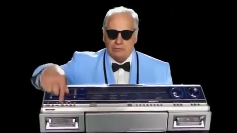 Lambnesia (2013), scene 4 at 00:29: Kekovich, now in a powder-blue tuxedo with a black bow tie and sunglasses, stands behind a large, retro boombox. He presses a button and is transported into a kaleidoscopic, psychedelic music video.