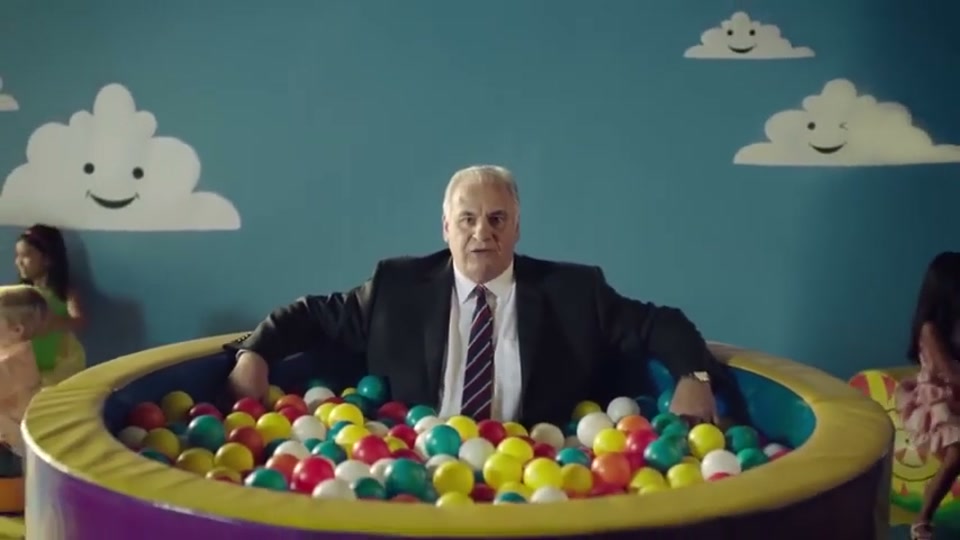 Generation Lamb (2014), scene 1 at 00:05: Sam Kekovich, a middle-aged man in a dark suit and tie, sits in a colourful children's ball pit against a cartoonish blue sky backdrop with smiling clouds. The camera zooms out to reveal the full setting.