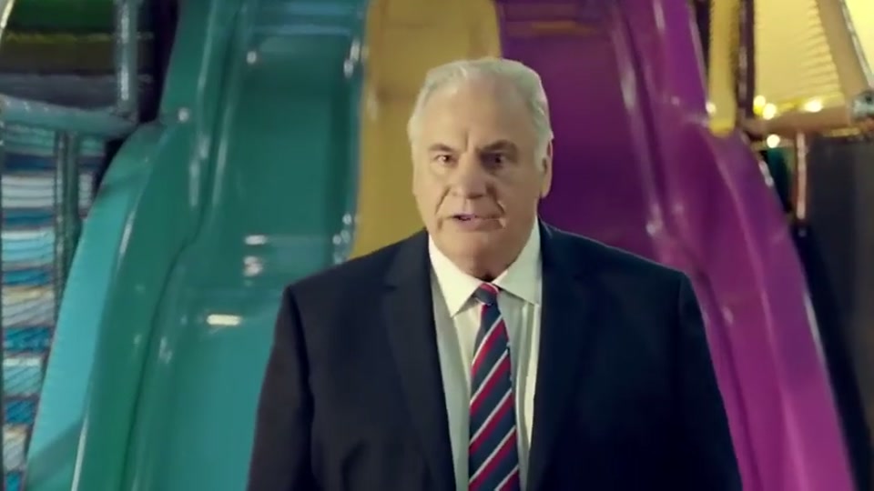 Generation Lamb (2014), scene 1 at 00:07: Sam Kekovich, a middle-aged man in a dark suit and tie, sits in a colourful children's ball pit against a cartoonish blue sky backdrop with smiling clouds. The camera zooms out to reveal the full setting.