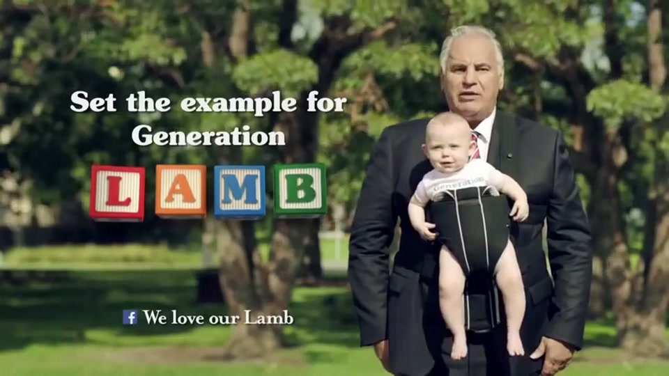Generation Lamb (2014), scene 6 at 00:57: Kekovich stands facing the camera, now wearing the baby from the previous scene in a front-facing baby carrier. The on-screen text appears next to him.