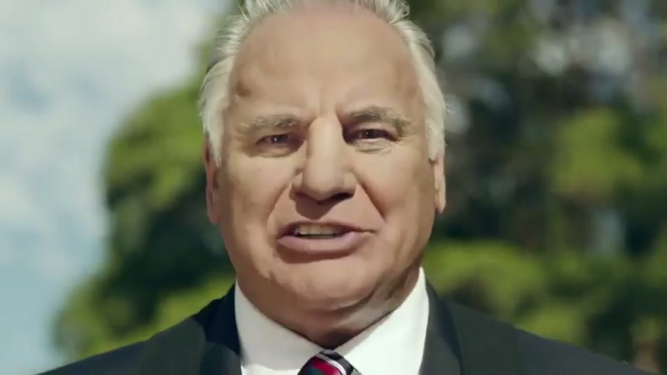 Generation Lamb (2014), scene 6 at 00:55: Kekovich stands facing the camera, now wearing the baby from the previous scene in a front-facing baby carrier. The on-screen text appears next to him.