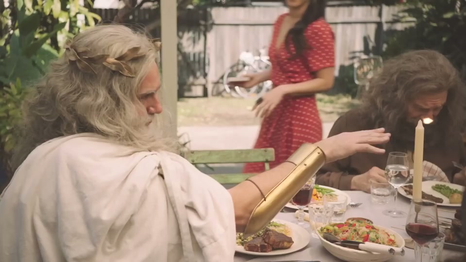 Gods (You Never Lamb Alone) (2017), scene 1 at 00:09: The scene opens on a long dinner table in a brightly lit backyard patio. A man dressed as the Greek god Zeus, with a white beard, toga, and golden laurel wreath, is speaking to a man dressed as Jesus.