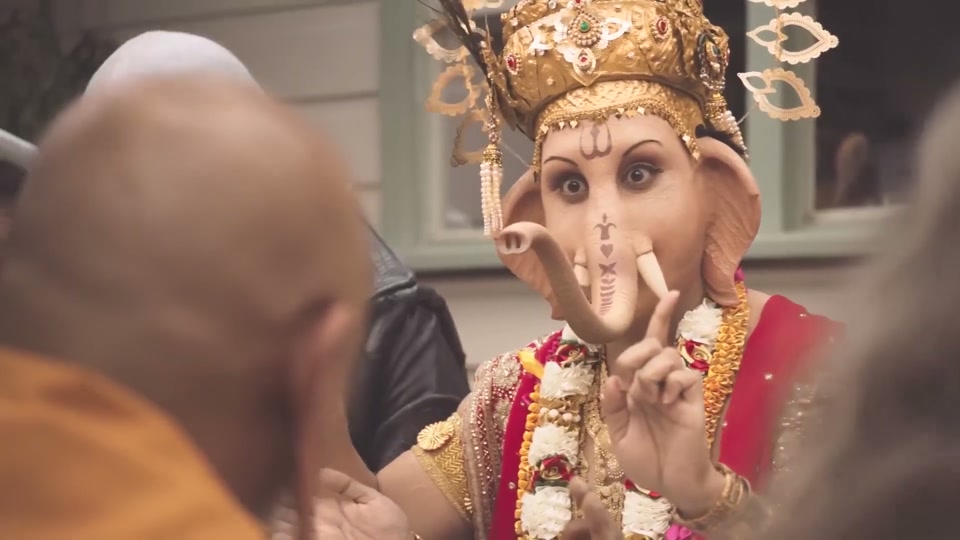 Gods (You Never Lamb Alone) (2017), scene 4 at 00:46: A man dressed as Buddha speaks to the table. The camera focuses on Ganesha, who is wearing ornate Hindu attire and has an elephant head. Ganesha responds dryly. Buddha laughs.