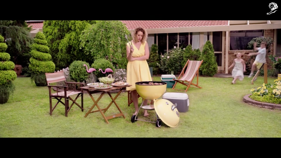 Lamb Side Story (2018), scene 1 at 00:05: The scene opens with an overhead shot of a quiet, manicured suburban cul-de-sac. A woman in a bright yellow dress is in her backyard, tending a matching yellow kettle barbecue filled with sizzling lamb chops and kebabs.