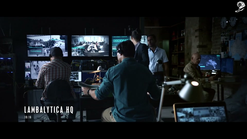 Lambalytica (2020), scene 2 at 00:19: Inside the dark, server-filled headquarters of "Lambalytica," agents monitor screens showing data maps of Australia and CCTV feeds. A stern female executive explains the operation to a new recruit.