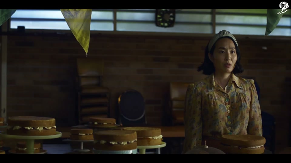 Un-Australian (2023), scene 1 at 00:24: The scene is a dimly lit community hall, set up for a baking competition at an "Annual Show." A stern, older female judge inspects several identical sponge cakes. She pins a first-place ribbon on one.