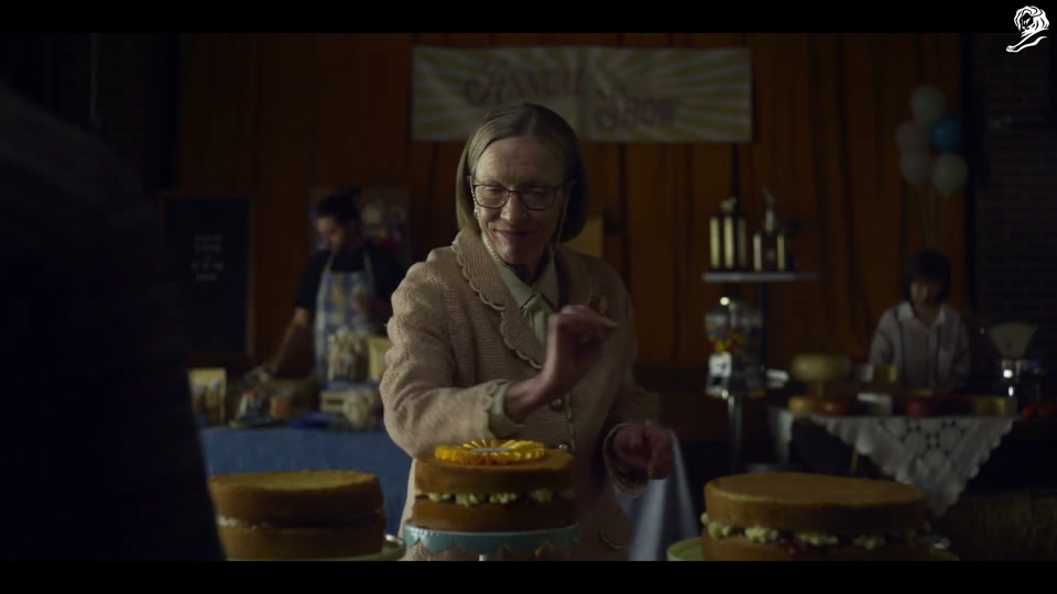 Un-Australian (2023), scene 1 at 00:08: The scene is a dimly lit community hall, set up for a baking competition at an "Annual Show." A stern, older female judge inspects several identical sponge cakes. She pins a first-place ribbon on one.
