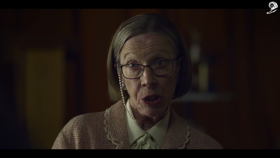 Un-Australian (2023), scene 1 at 00:16: The scene is a dimly lit community hall, set up for a baking competition at an "Annual Show." A stern, older female judge inspects several identical sponge cakes. She pins a first-place ribbon on one.