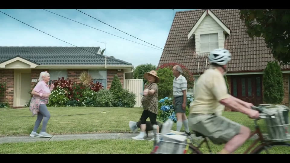 The Generation Gap (2024), scene 1 at 00:12: The scene opens in a quiet, manicured Australian suburb called "Boomertown." An elderly man on a bicycle, wearing a helmet, does a paper round. He throws a newspaper onto a perfect green lawn.