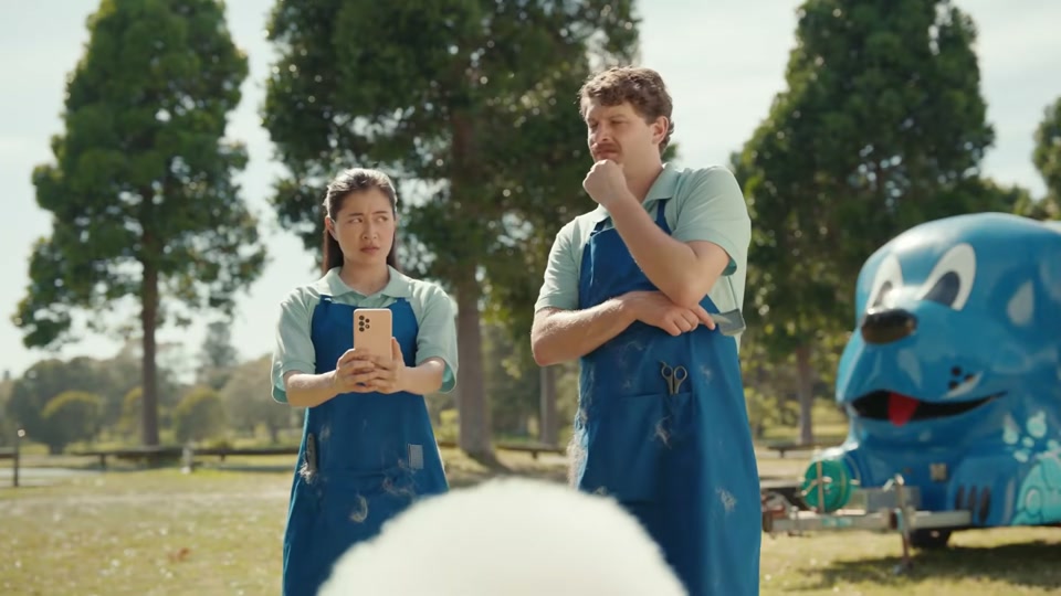 The Comments Section (2025), scene 1 at 00:04: Two professional dog groomers, a man and a woman, stand in a park admiring their work. They are dressed in matching light green polo shirts and blue aprons.