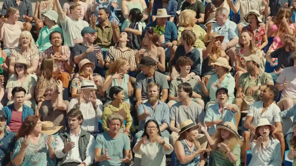 The Comments Section (2025), scene 2 at 00:24: The groomers look up, startled. The camera pulls back to reveal they are now standing on a stage before a colossal stadium grandstand filled with thousands of people.