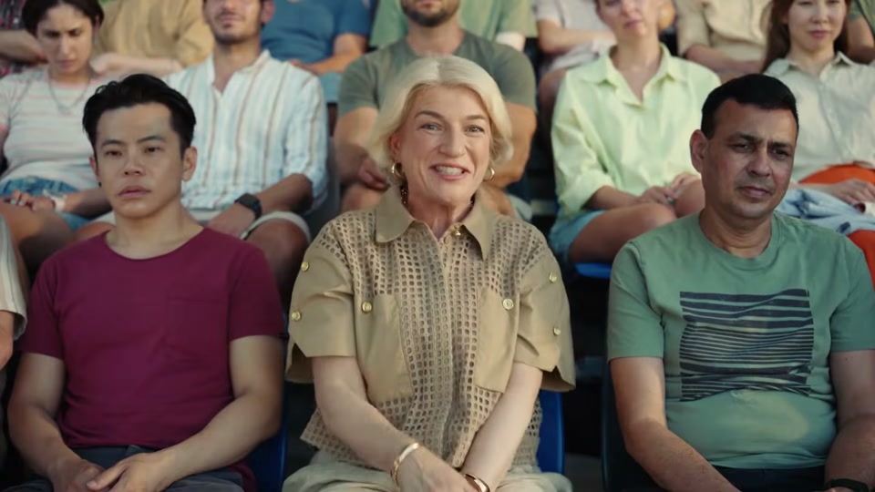 The Comments Section (2025), scene 2 at 00:13: The groomers look up, startled. The camera pulls back to reveal they are now standing on a stage before a colossal stadium grandstand filled with thousands of people.