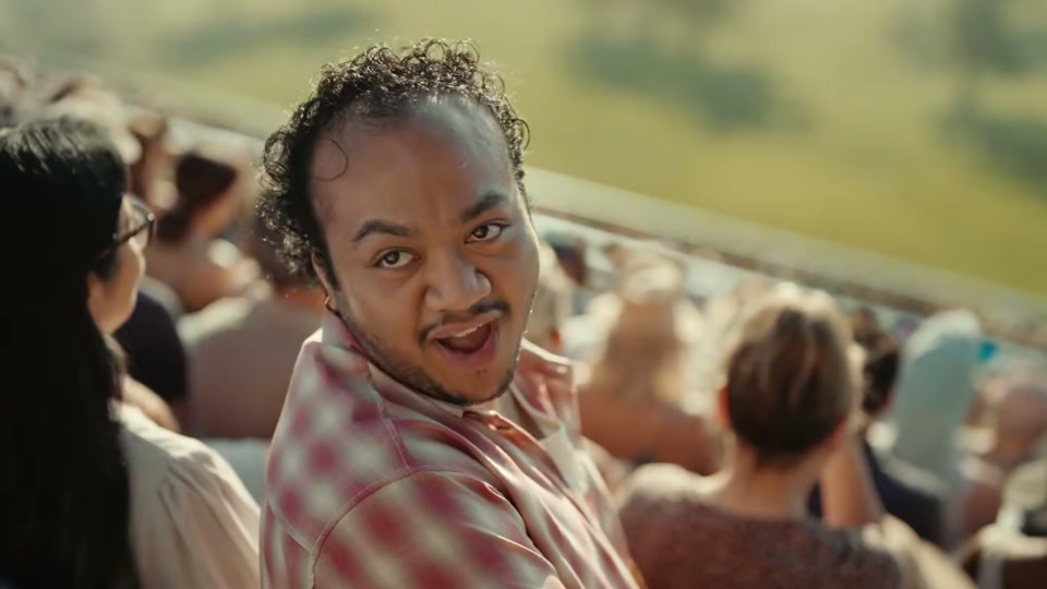 The Comments Section (2025), scene 2 at 00:19: The groomers look up, startled. The camera pulls back to reveal they are now standing on a stage before a colossal stadium grandstand filled with thousands of people.