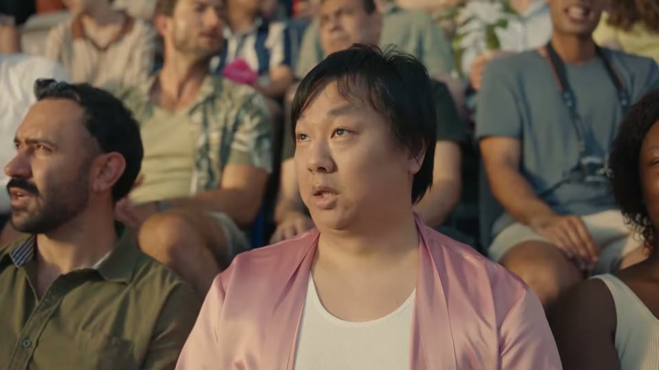 The Comments Section (2025), scene 3 at 00:51: The groomers look up at the sky, concerned. The scene shifts to an aerial shot of a wind turbine on a rolling green hill by the sea. The entire stadium grandstand is now superimposed on the hill.