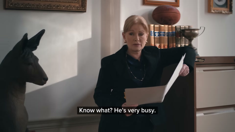 Happiness Index (2026), scene 1 at 00:10: The scene opens in a stately, wood-panelled office. Three large windows look out onto a tree-lined street. An older man in a suit, Sam Kekovich, sits at a large wooden desk, looking sternly at paperwork.