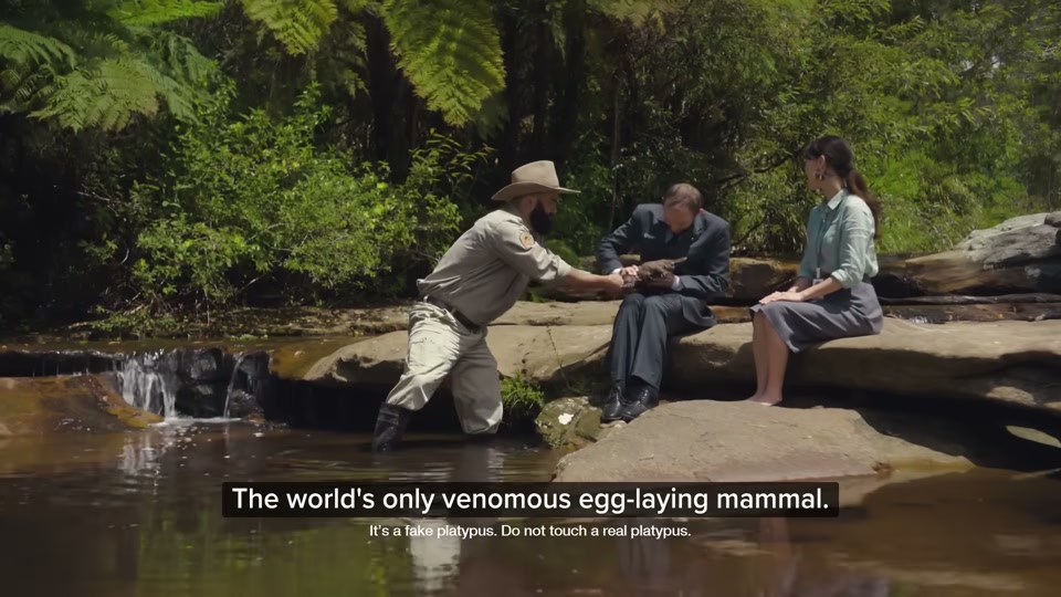 Happiness Index (2026), scene 4 at 01:12: The group is now in the back of the car, driving through a city. They then drive through a lush, green forest. Next, they are by a creek, where a park ranger is showing a male auditor a platypus.