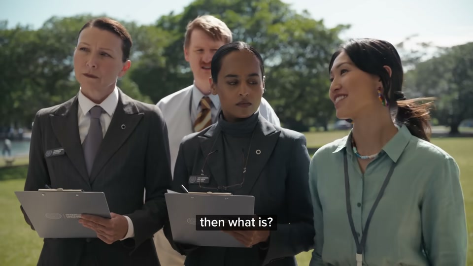Happiness Index (2026), scene 6 at 02:23: The group arrives at a large, sunny park by the water, filled with a diverse crowd of people at a massive public barbecue. Sam Kekovich is at the centre, presiding over a large public grill laden with lamb.