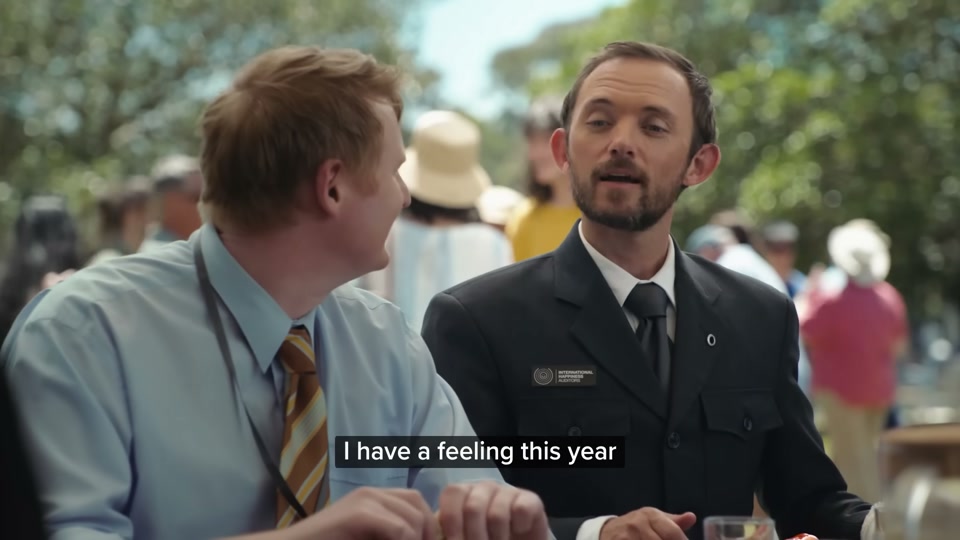 Happiness Index (2026), scene 6 at 02:49: The group arrives at a large, sunny park by the water, filled with a diverse crowd of people at a massive public barbecue. Sam Kekovich is at the centre, presiding over a large public grill laden with lamb.